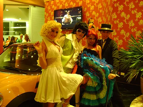Actores caracterizados como Marilyn Monroe y Elvis Presley junto a un carro amarillo