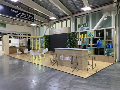 Stand ferial de Uniban en el Pabellón Azul de Plaza Mayor Medellín, con mostrador de madera, mesas altas, banquetas y mobiliario blanco